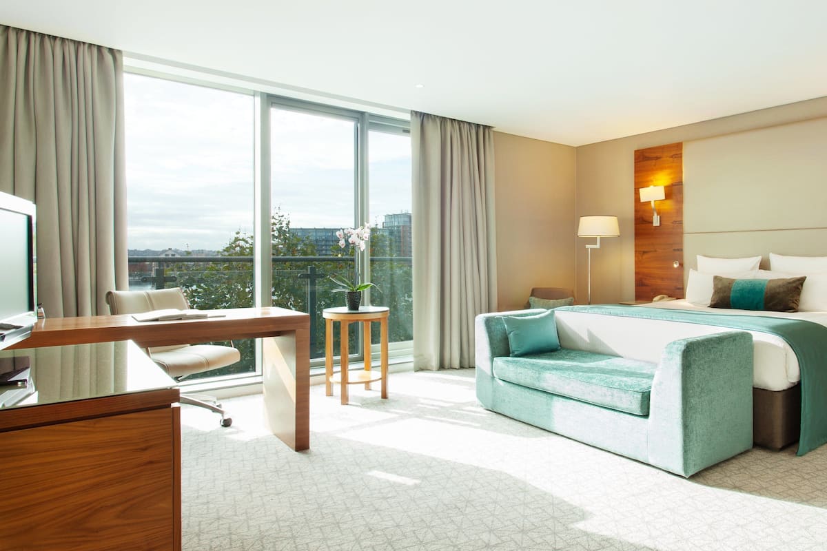 Een moderne hotelkamer met een groot bed, een bureau en een stoel. Er is een raam met gordijnen, een lamp en een klein tafeltje met een plant.