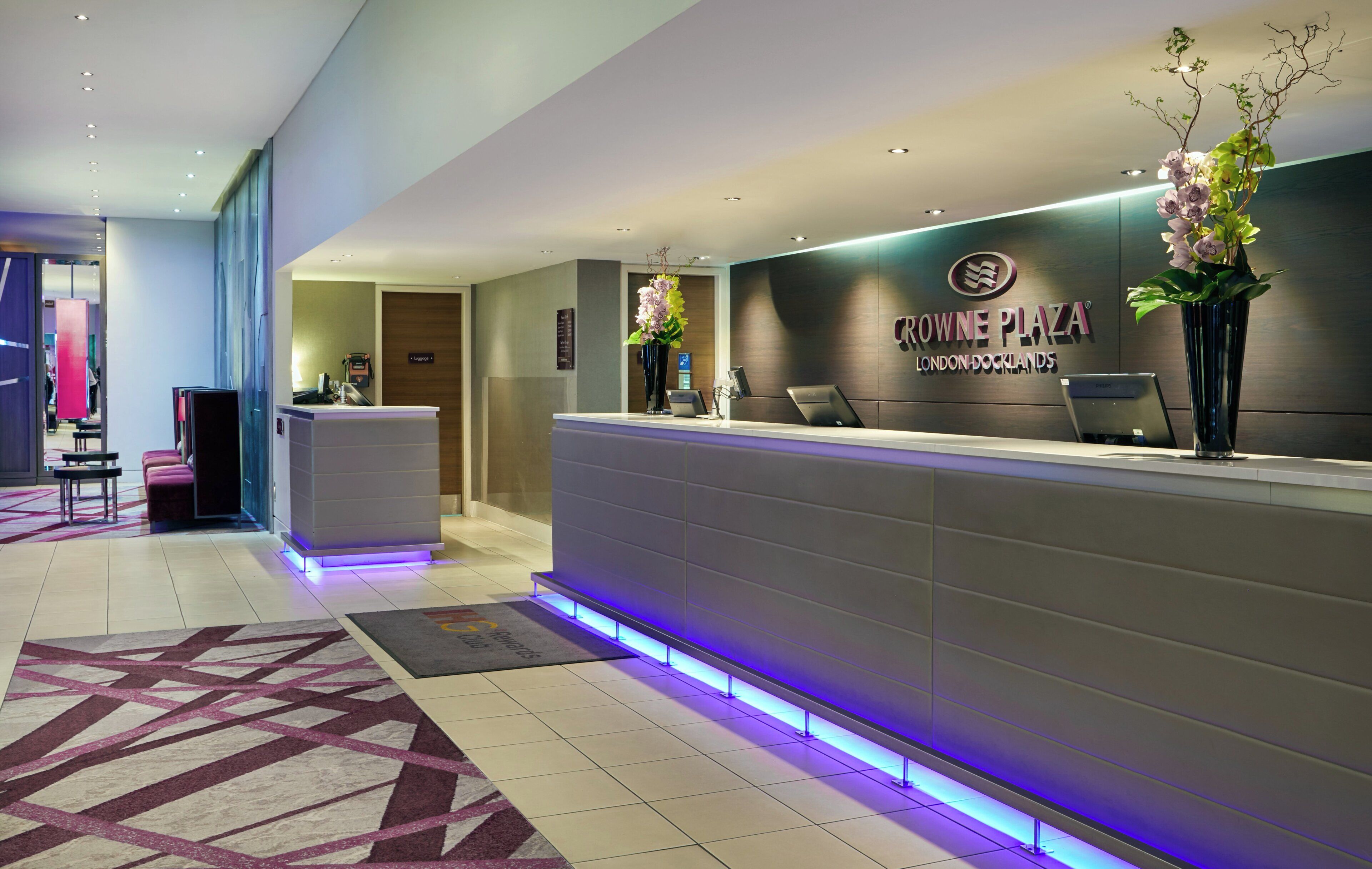 A modern reception desk with a sign for Crowne Plaza London Docklands, a luggage area, and a reception desk with a computer and a vase of flowers.