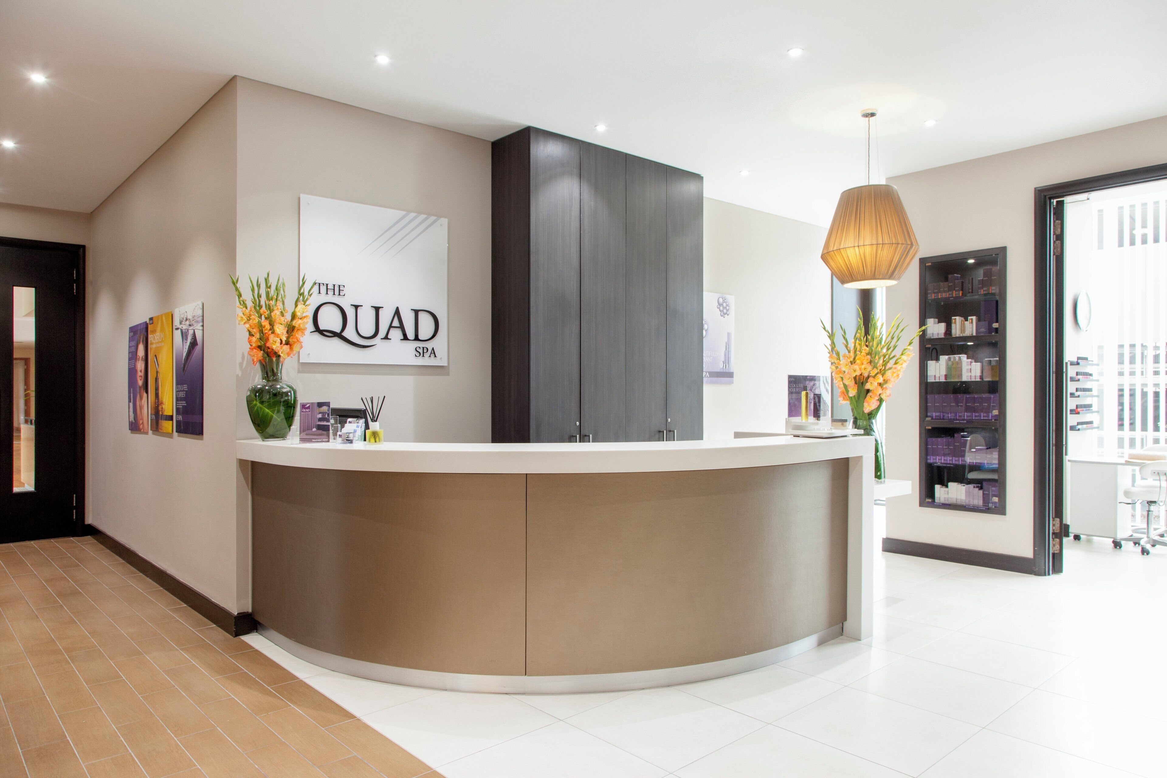 A reception desk with a sign reading 'The Quad SPA', a modern interior with a large cabinet, a hanging lamp, and a bookshelf with various items.