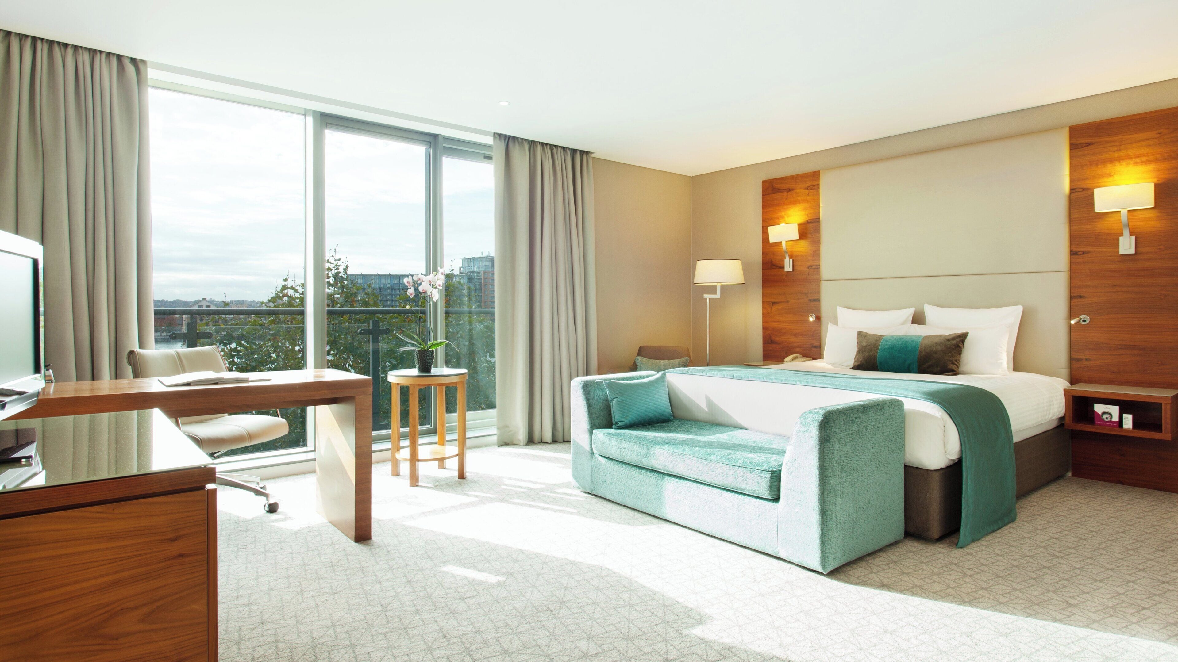 A hotel room with a bed, desk, chair, and a view of the city.