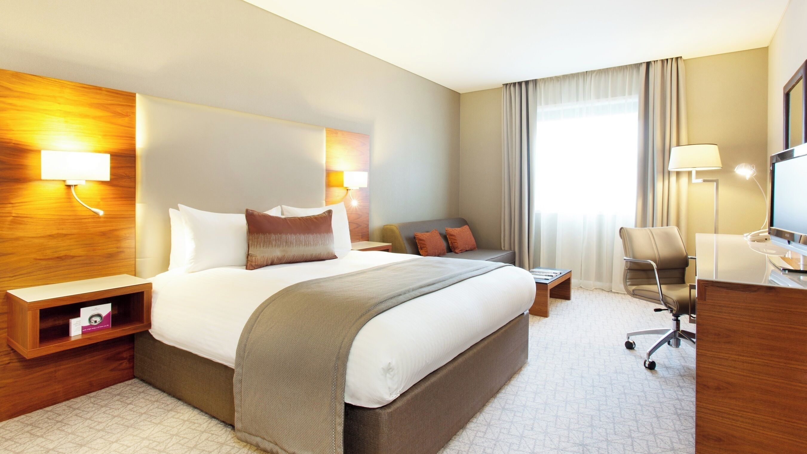 A hotel room with a large bed, bedside tables, a desk, a chair, and a television.