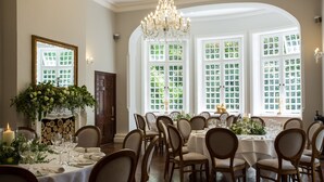 Un comedor formal con varias mesas dispuestas para una comida, amplias ventanas con follaje verde y un candelabro.
