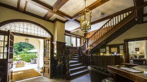 Una imponente escalera de madera con barandales y un candelabro, que conduce a un área de recepción con un mostrador y asientos.