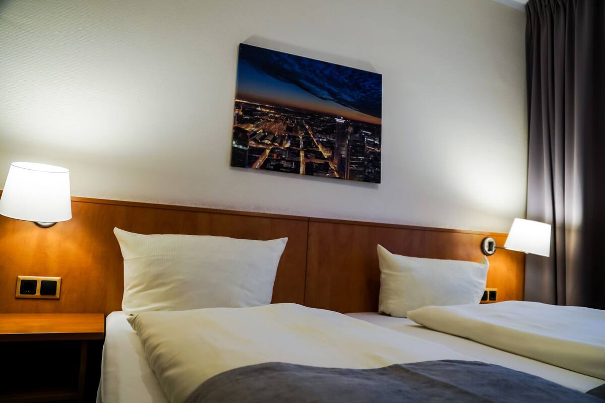 Habitación de hotel con cama, dos lámparas de noche, un cuadro de un paisaje urbano nocturno en la pared y una ventana con cortinas.