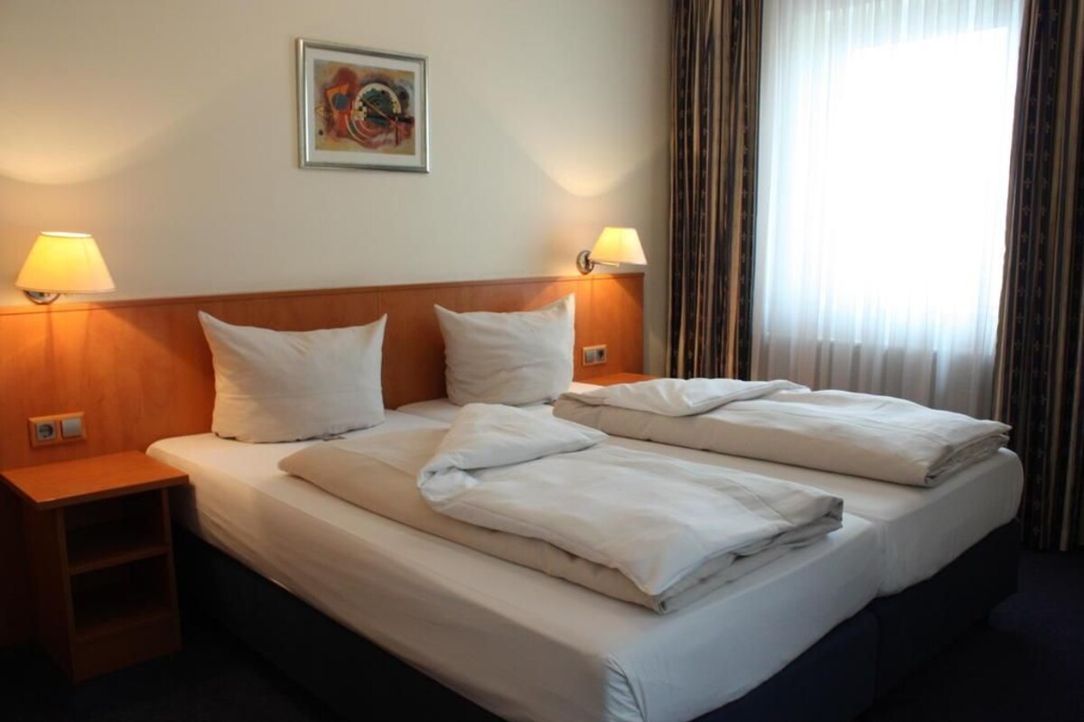 A hotel room with a bed, two bedside lamps, a nightstand, a wall-mounted painting, and a window with curtains.