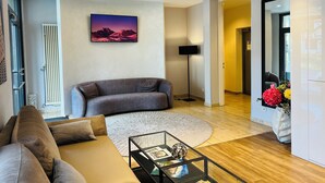 Een moderne woonkamer met een grijze bank, een salontafel met tijdschriften, een vloerlamp en een schilderij van bergen aan de muur.
