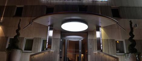 La entrada al hotel Niederräder Hof, con un diseño moderno, presenta una fachada curva, un llamativo letrero iluminado y una escalera que conduce a la entrada.