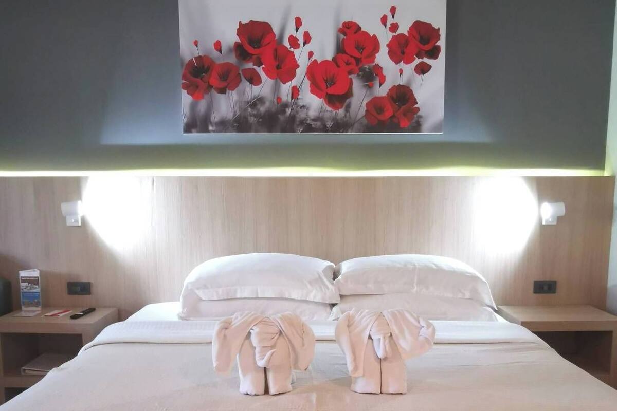 Ein Hotelzimmer mit einem Bett, zwei Nachttischen, einem Bild von roten Blumen und zwei Elefanten-Formen aus Handtüchern auf dem Bett.