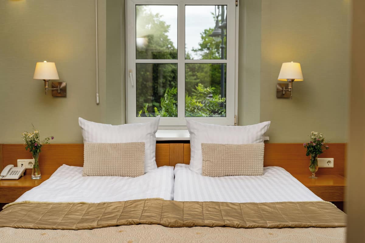 A double bed with a beige bedspread and two pillows, a bedside table with a lamp, a vase with flowers, and a window with a view of greenery.