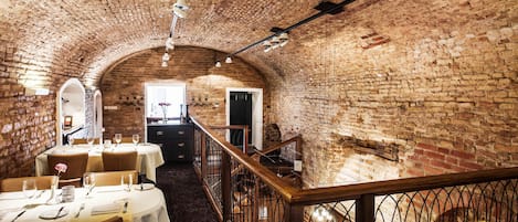 A dining area with brick walls, white tables, and chairs. There is a staircase with a railing leading to an upper level.