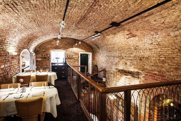 A dining area with brick walls, white tables, and chairs. There is a staircase with a railing leading to an upper level. - Mabre Residence Hotel (Vilnius)