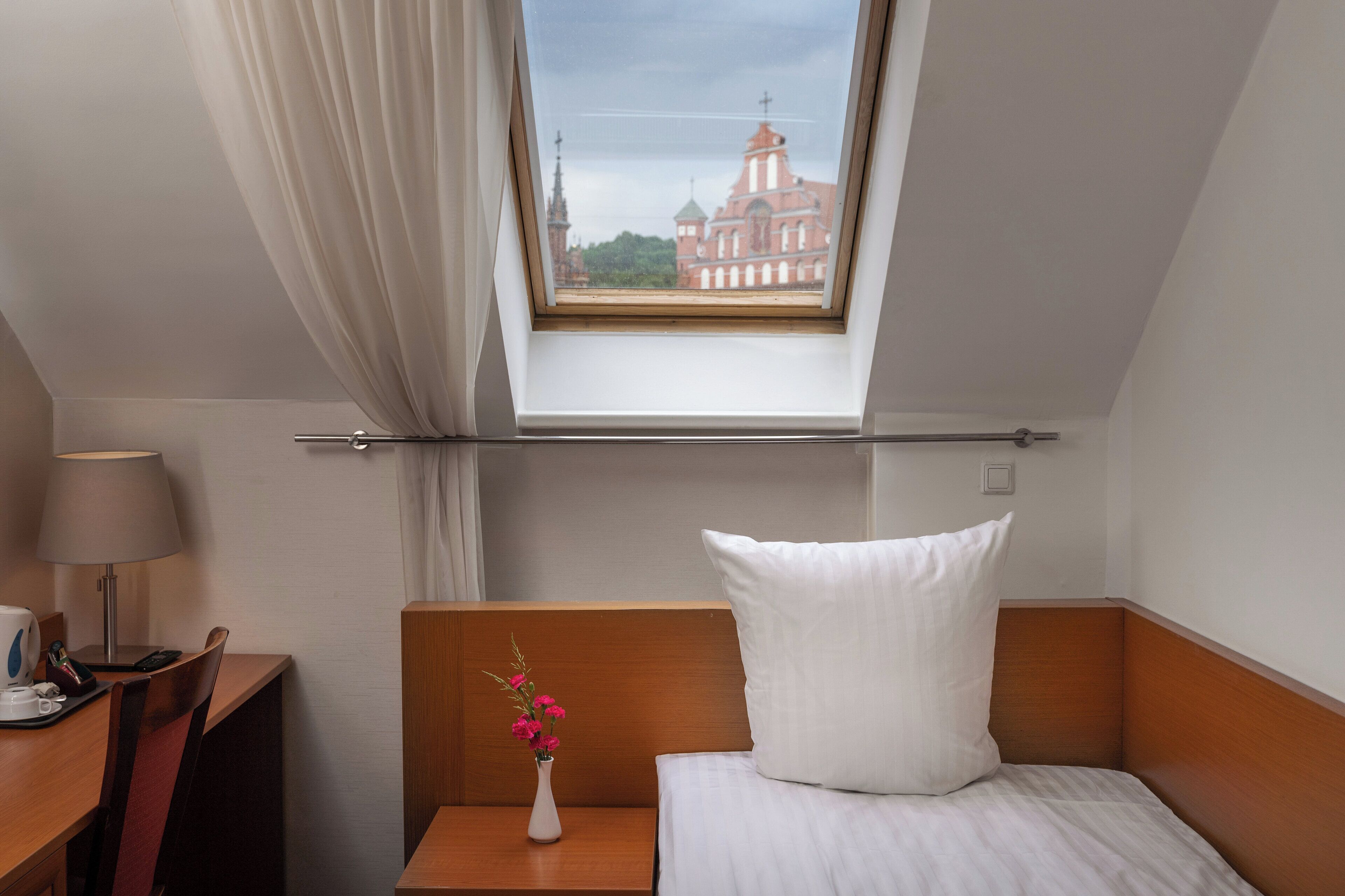 A hotel room with a skylight, a bed, a nightstand, a desk, a lamp, and a vase with flowers.