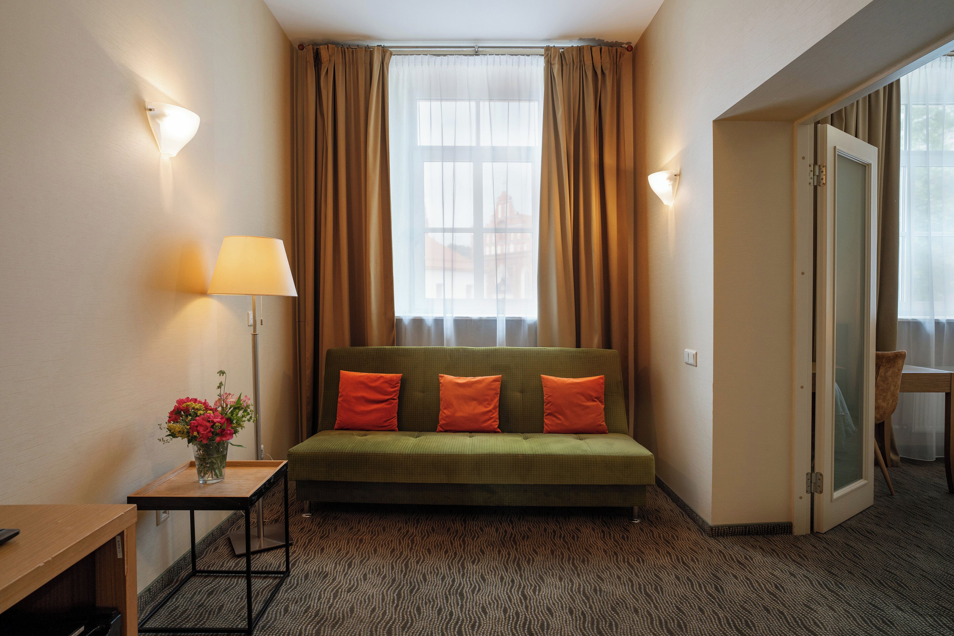 A hotel room with a green sofa, two orange pillows, a small table with a lamp, and a vase of flowers.