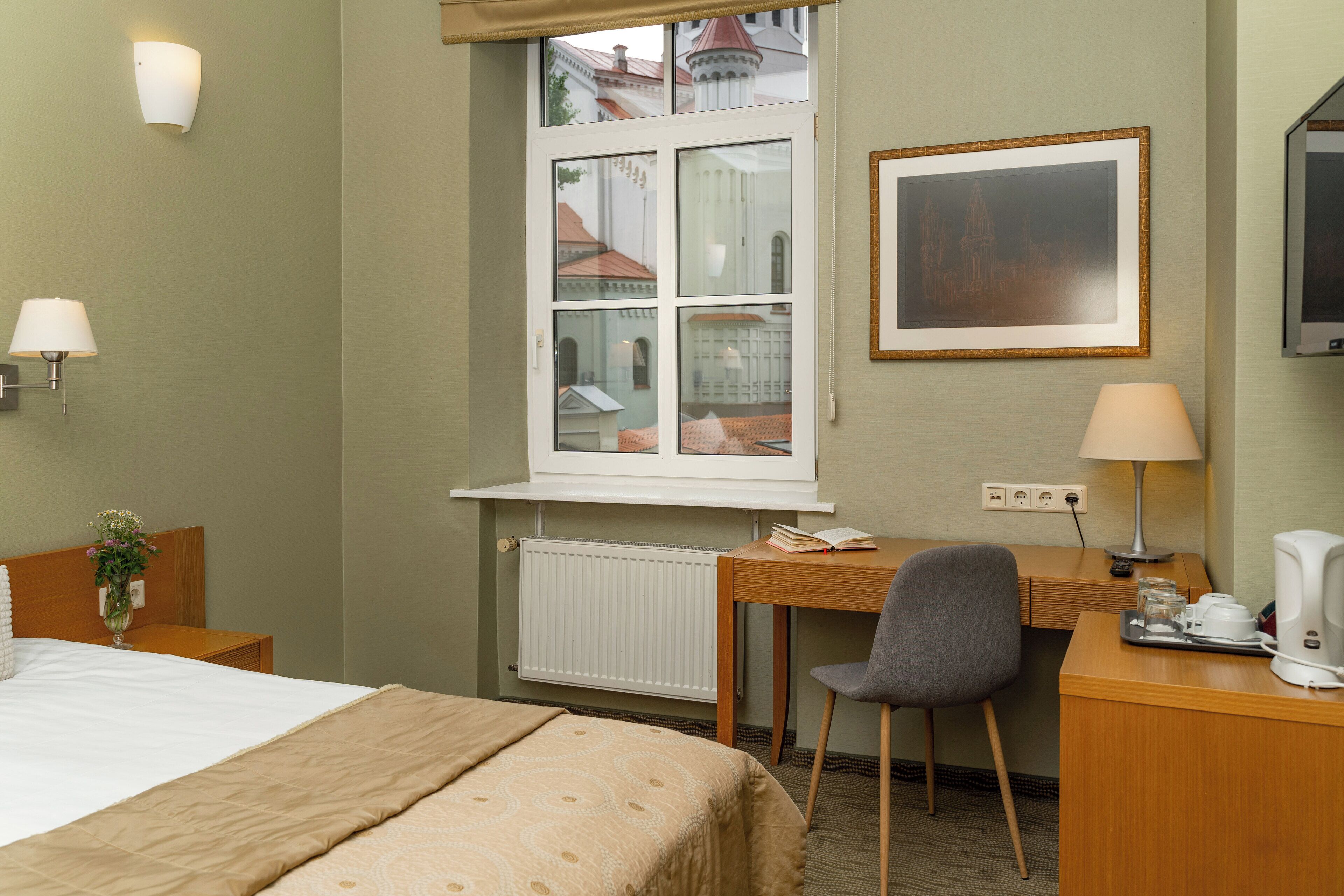 A hotel room with a bed, a desk, a chair, a window with a view of buildings, a framed picture on the wall, and a television.