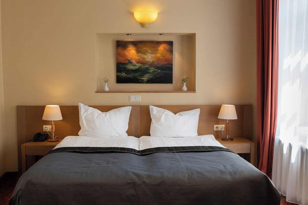 A hotel room with a bed, two bedside tables with lamps, a painting on the wall, and a window with curtains.