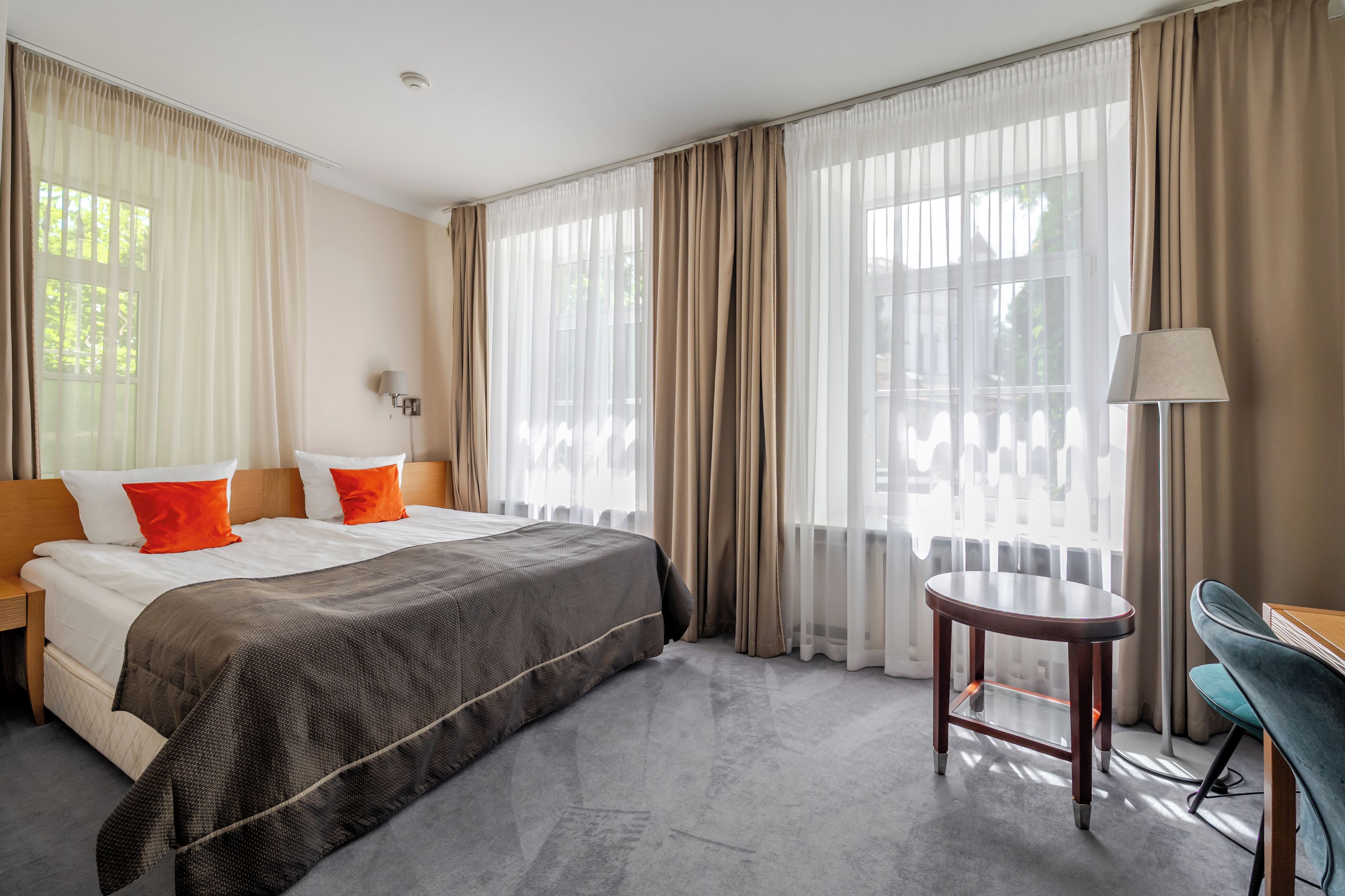A hotel room with a bed, a nightstand, a lamp, a chair, and large windows with curtains.