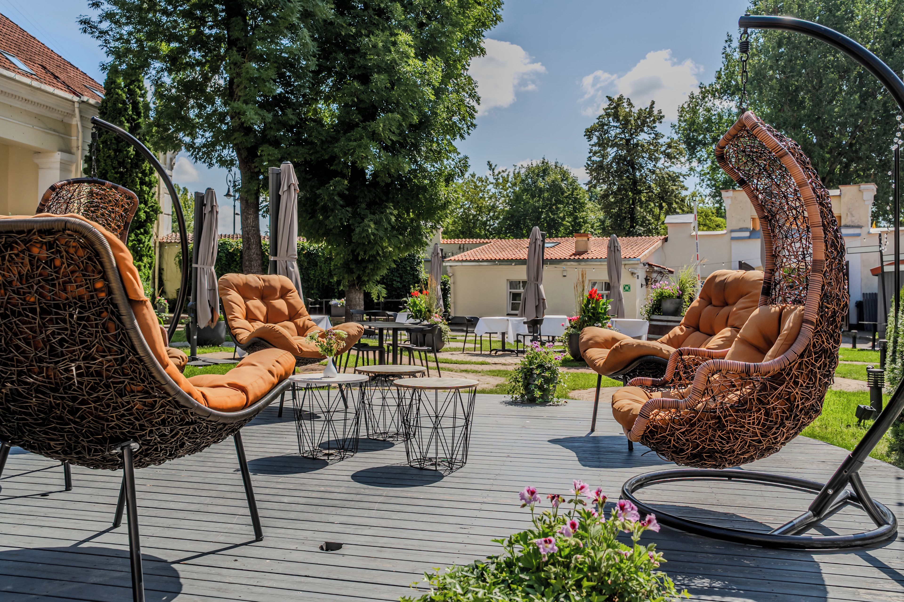 An outdoor seating area with wicker chairs and a hanging chair, surrounded by greenery and a building with a tiled roof.