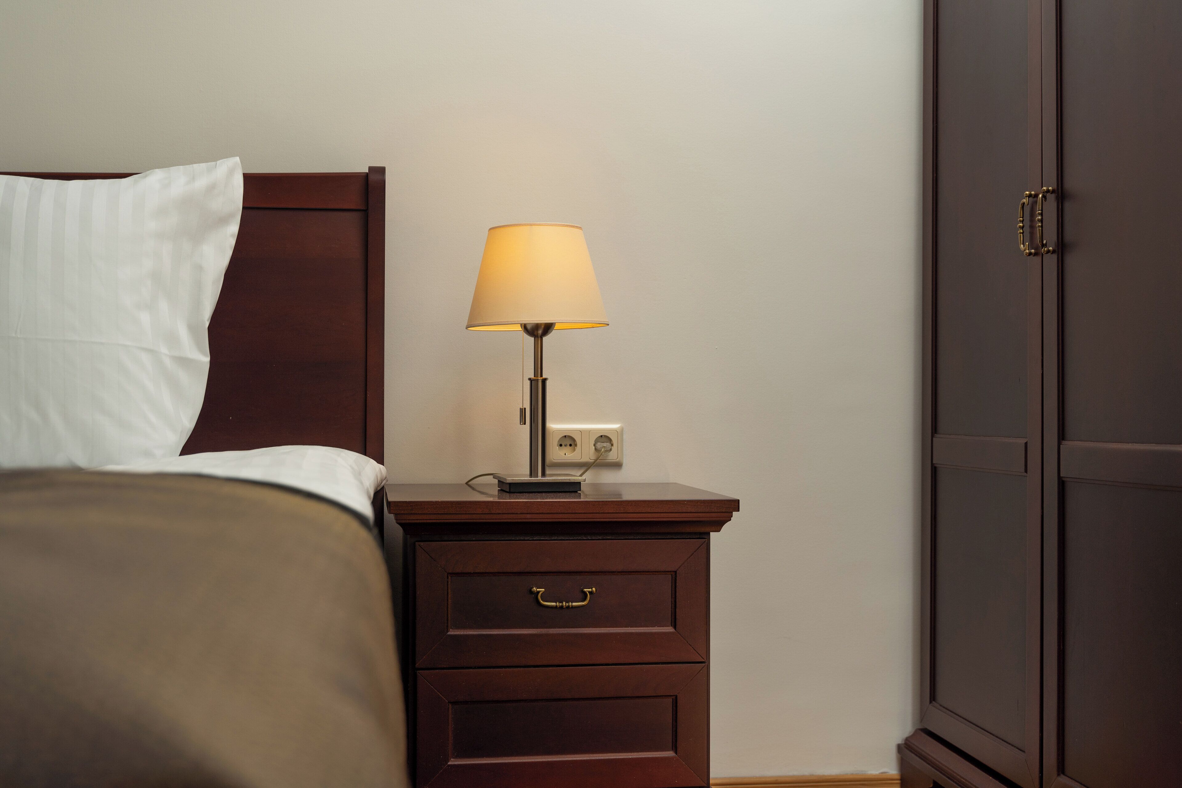 A neatly made bed with a white pillow and a brown blanket, a bedside table with a lamp, and a dark wooden wardrobe.