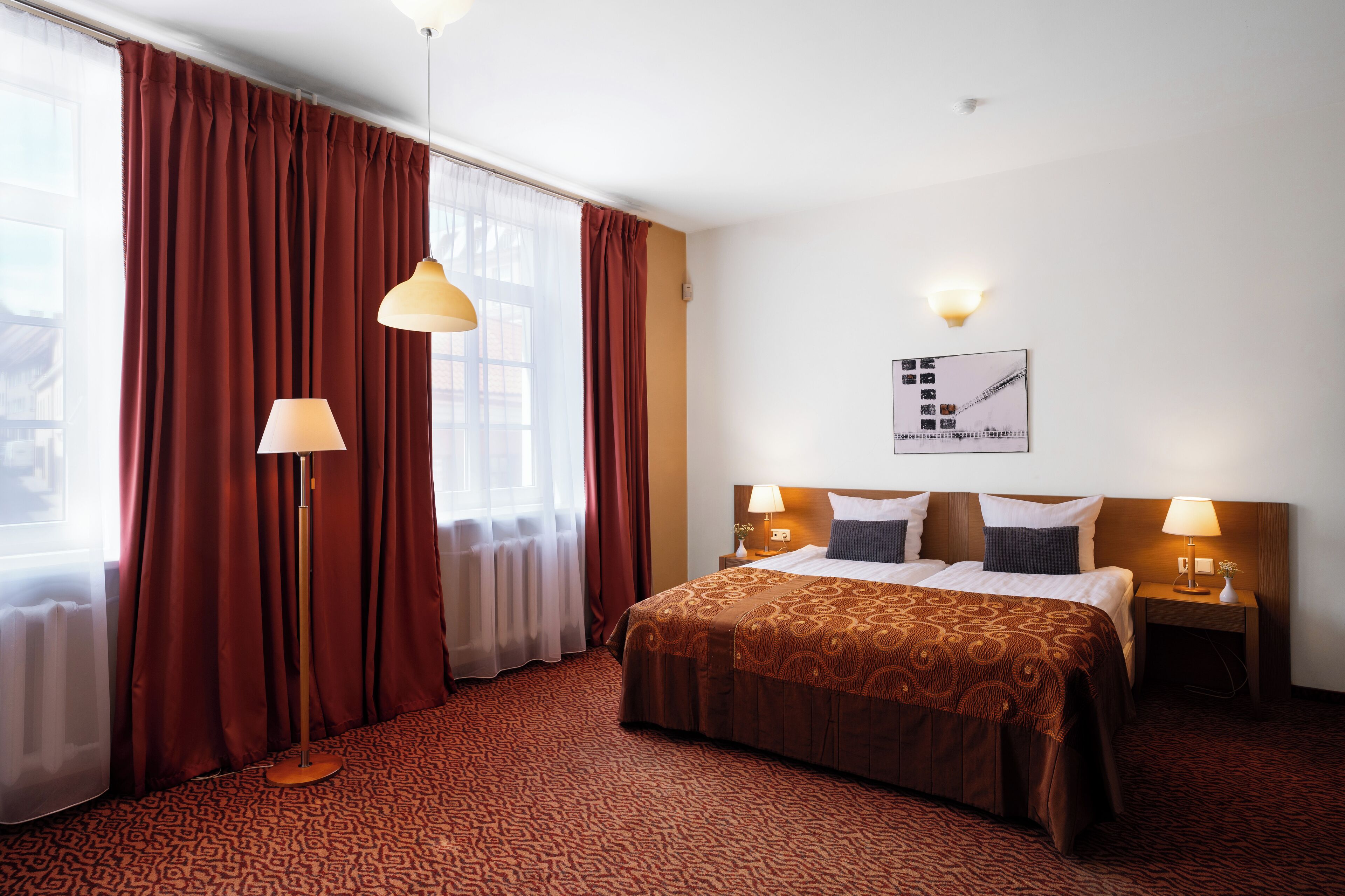 Habitación de hotel con una cama grande, dos mesitas de noche con lámparas, una ventana con cortinas y un cuadro colgado en la pared.