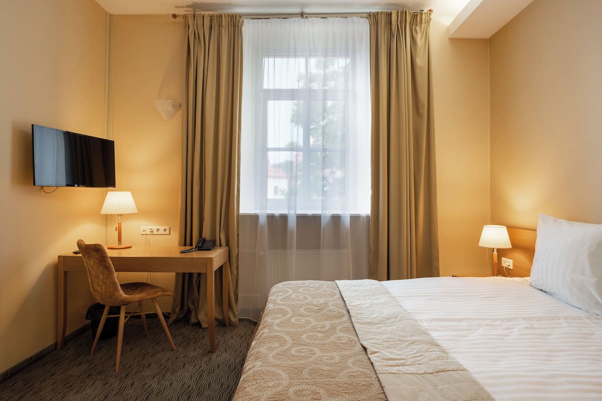 A hotel room with a bed, a desk with a chair, a television mounted on the wall, and a window with curtains.
