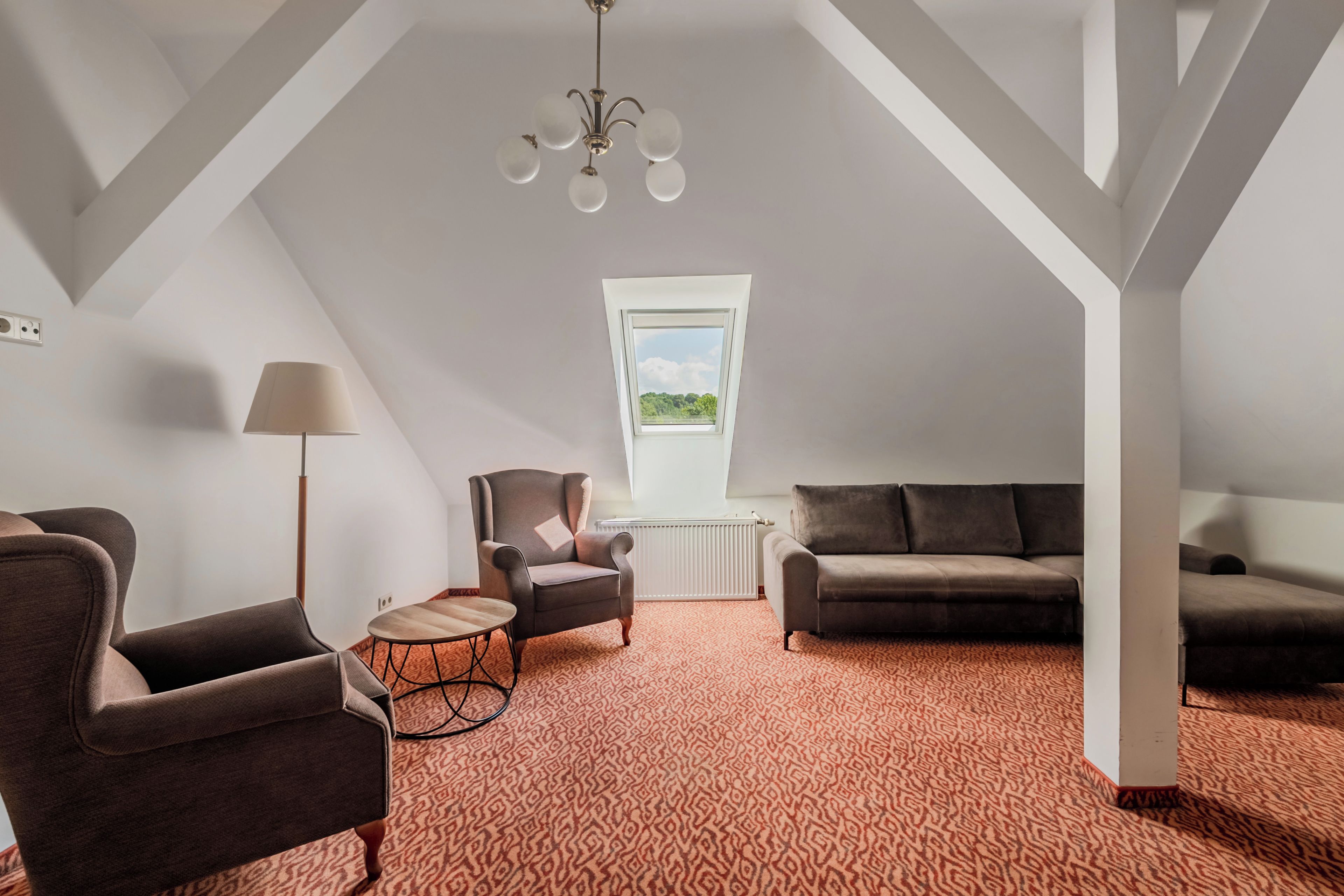 A spacious loft bedroom with a sloped ceiling, a large window, a chandelier, and a patterned carpet.