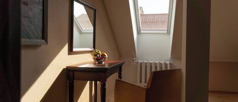 A small room with a sloped ceiling, a window, a radiator, a table with a fruit bowl, a chair, and a mirror on the wall.