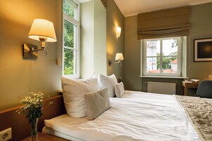 Una habitación de hotel con una cama grande, dos lámparas fijadas a la pared, un ventanal con vistas a edificios y una mesita con un libro.