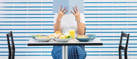 A person seated at a table with a meal, facing a window with horizontal blinds.