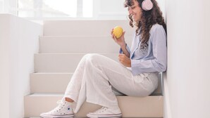 Una persona sentada en una escalera en una habitación bien iluminada, con un ventanal grande, usando audífonos y sosteniendo un limón.