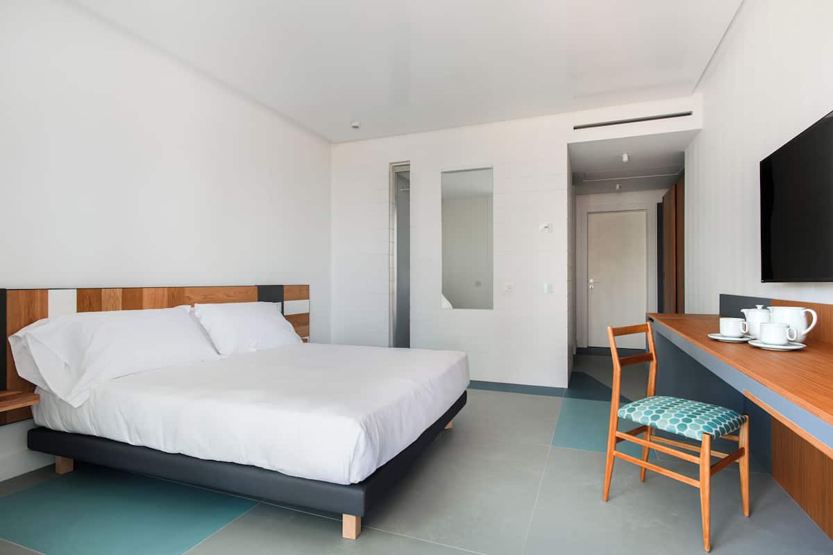 A modern hotel room with a bed, a wooden nightstand, a chair, a desk with a cup and saucer, and a large mirror on the wall.