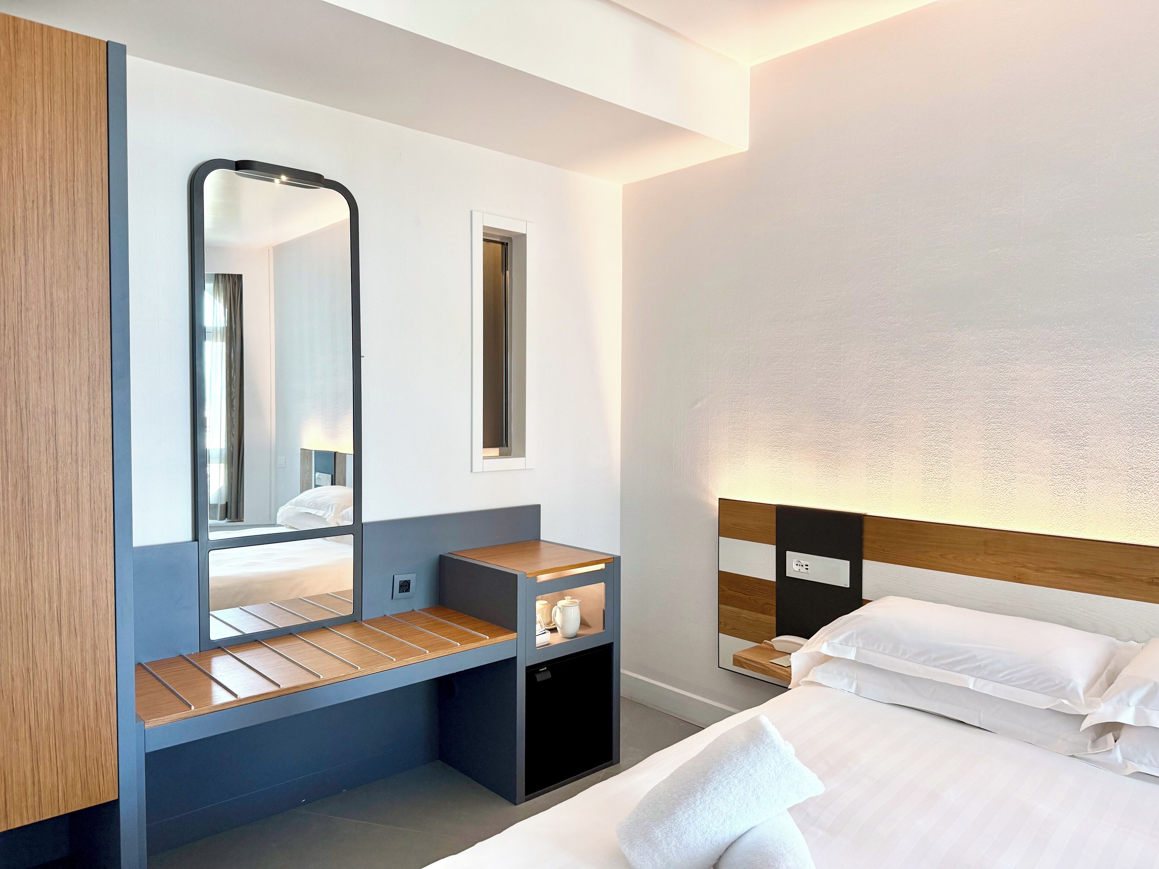 Une chambre d’hôtel moderne équipée d’un lit, d’une table de chevet, d’un miroir, d’un petit bureau et d’une lampe fixée au mur.