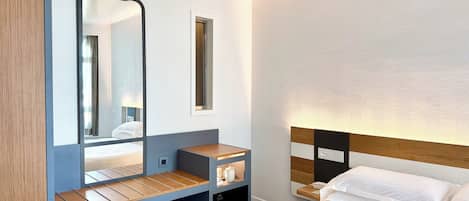 A modern hotel room with a bed, a bedside table, a mirror, a small desk, and a wall-mounted light.