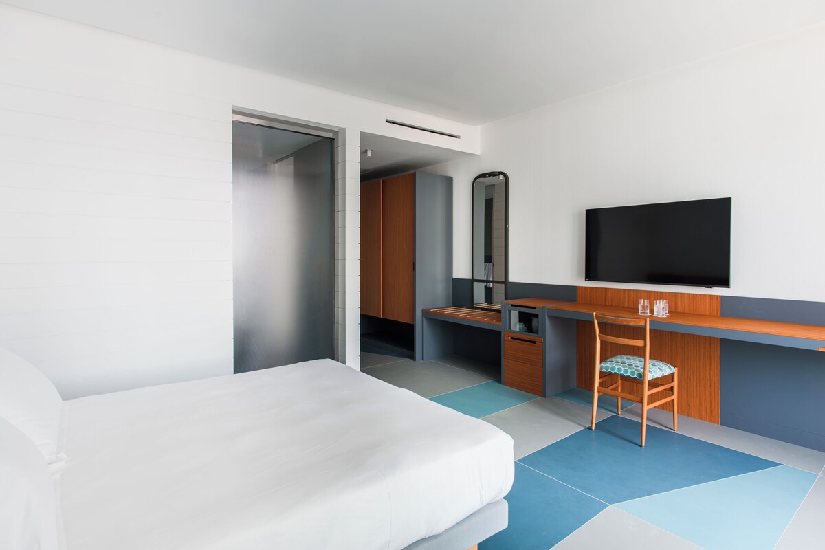 Una habitación de hotel moderna con cama, escritorio, silla, televisor y armario.