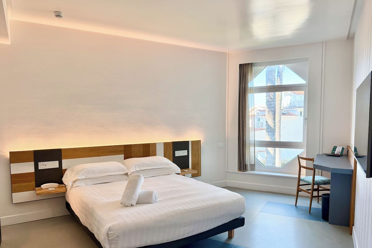 A modern hotel room with a large bed, bedside tables, a desk, and a window with a view.