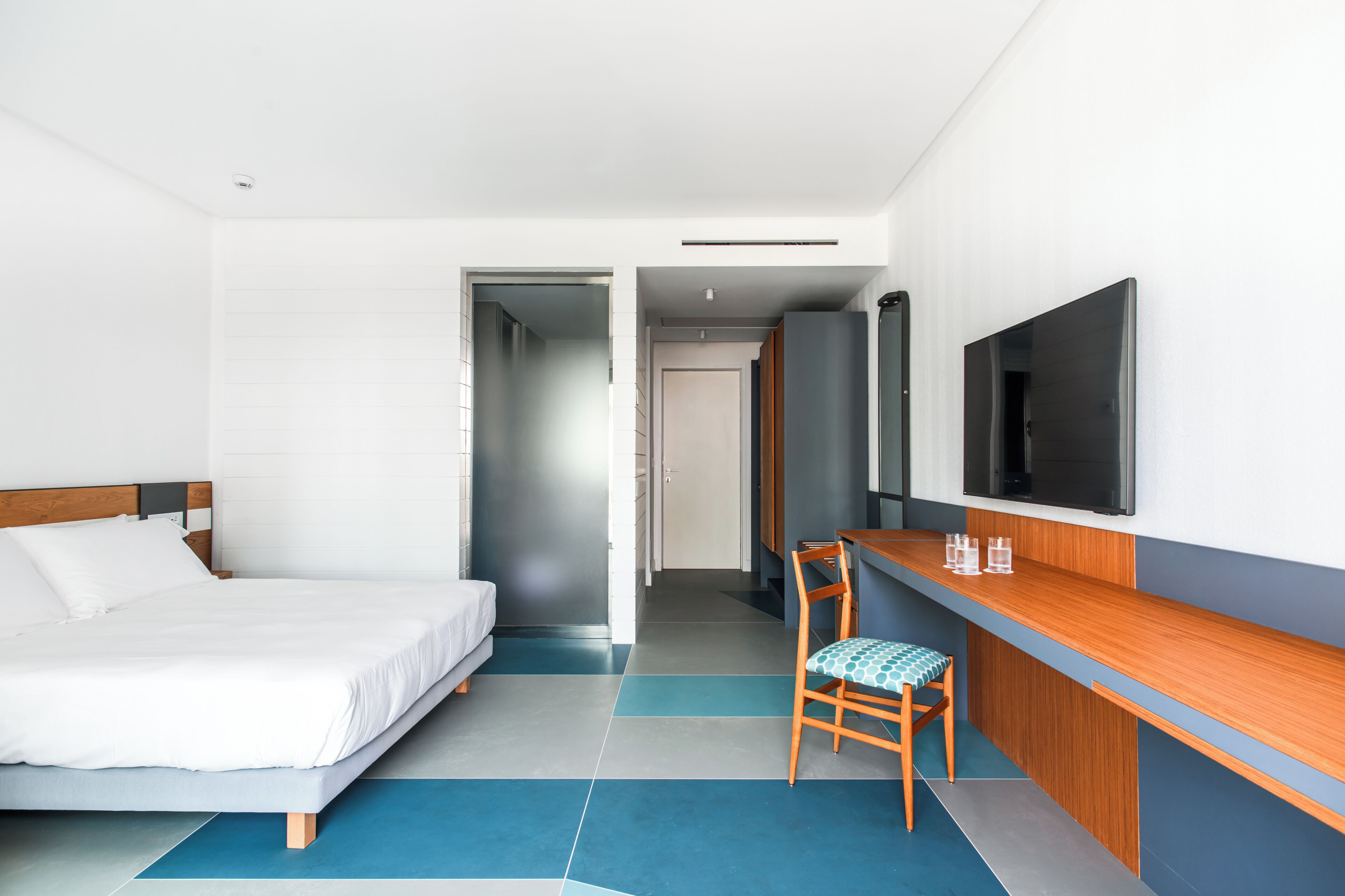 A hotel room with a bed, a desk with a chair, a television, and a door leading to another room.