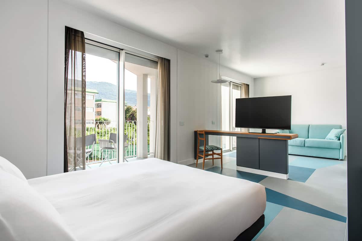 A modern hotel room with a large bed, a TV, a sofa, and a balcony with a view of the city.