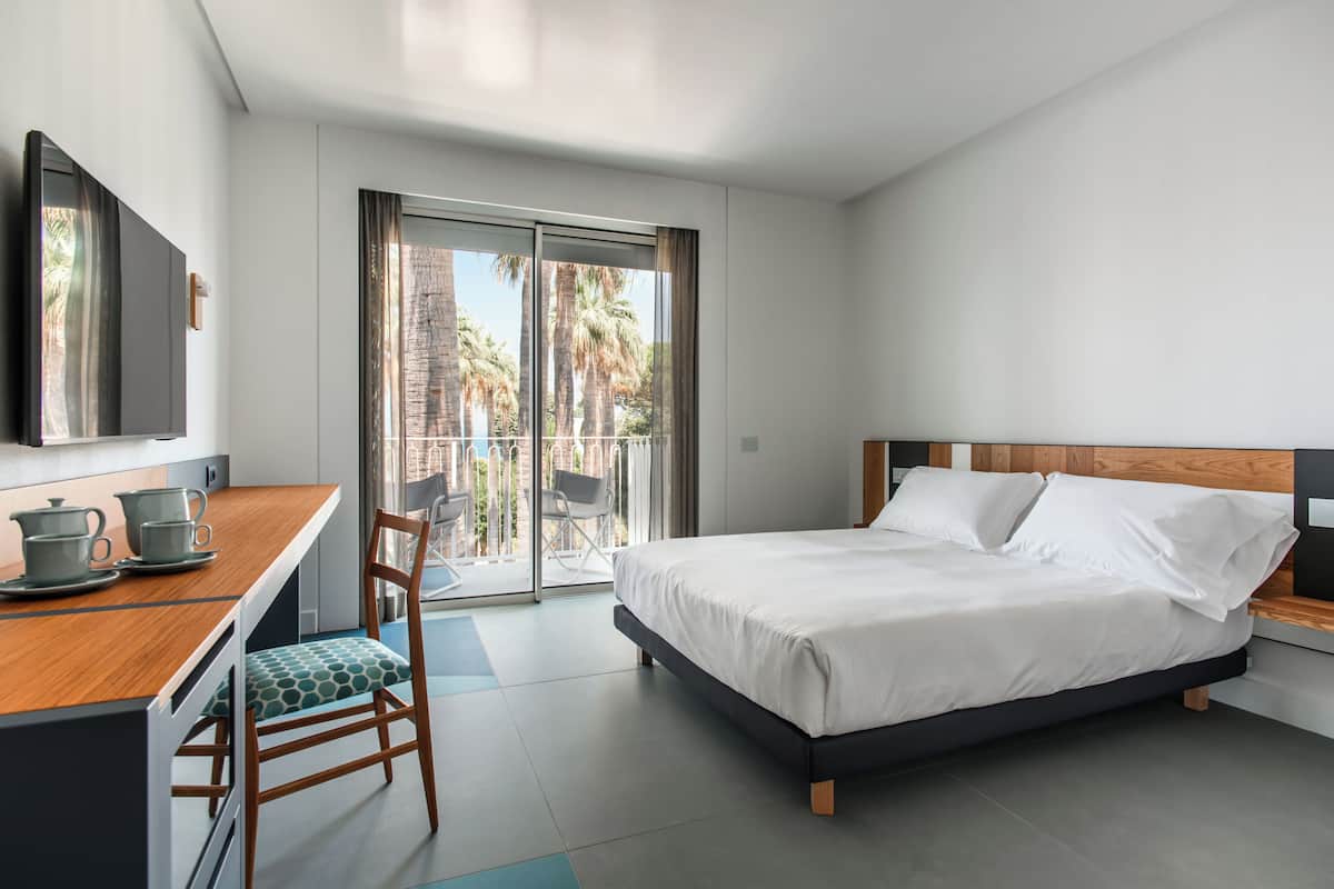 A modern hotel room with a bed, a desk with a chair, a television, and a balcony with a view of palm trees.