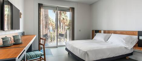 A modern hotel room with a bed, a desk with a chair, a television, and a balcony with a view of palm trees.