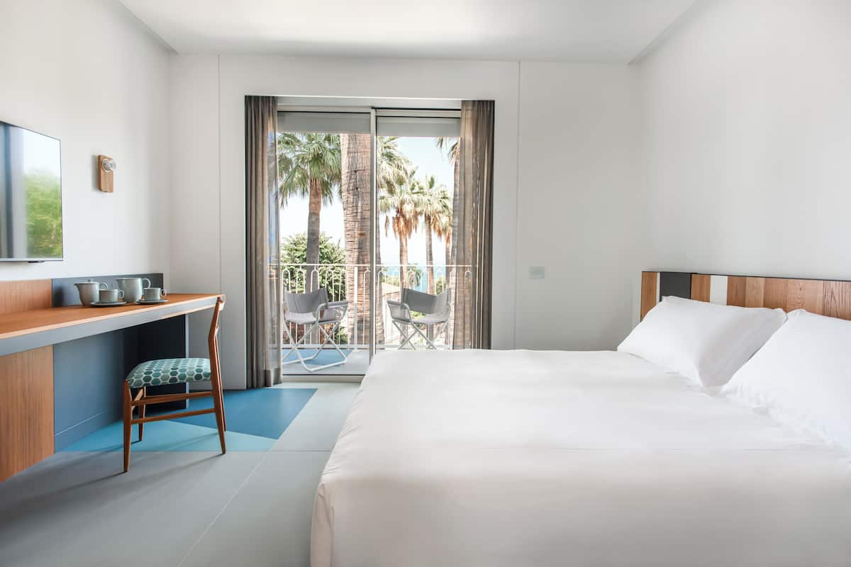 A hotel room with a bed, a desk with a chair, a TV, and a balcony with chairs.