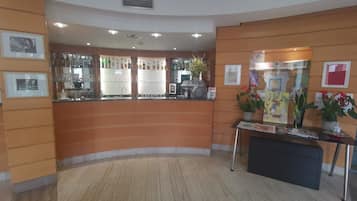 A reception area with a curved counter, a bar with various liquor bottles, framed artwork on the walls, and a small table with a newspaper and a potted plant.