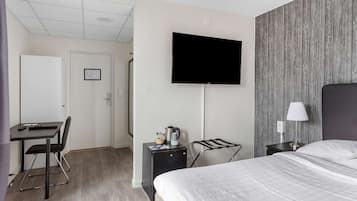 Ein Hotelzimmer mit einem Bett, einem Schreibtisch mit Stuhl, einem an der Wand befestigten Fernseher, einem kleinen Tisch mit Wasserkocher und einer Lampe auf einem Nachttisch.