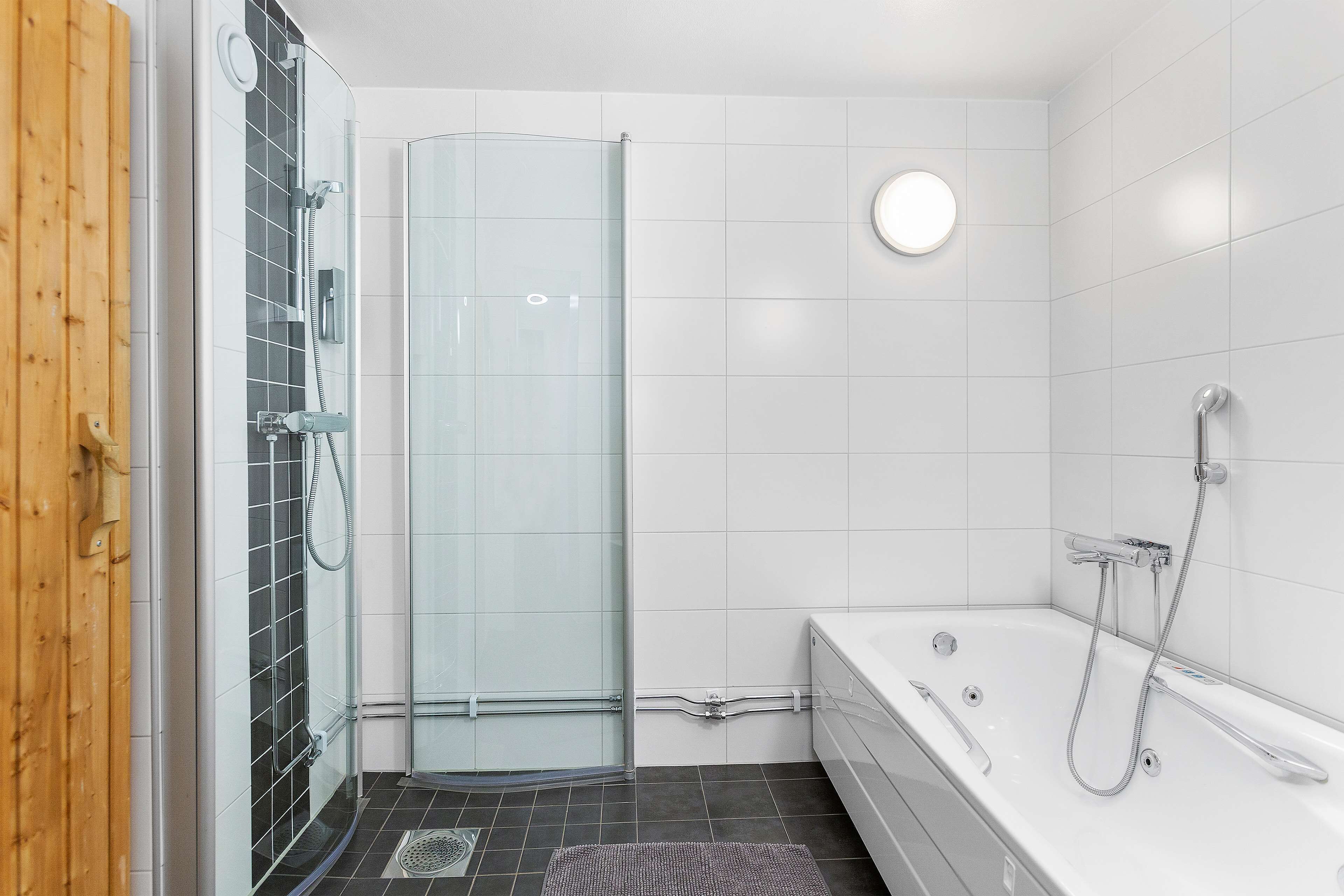 A modern bathroom with a glass shower enclosure, a bathtub with a handheld showerhead, and a wooden door.