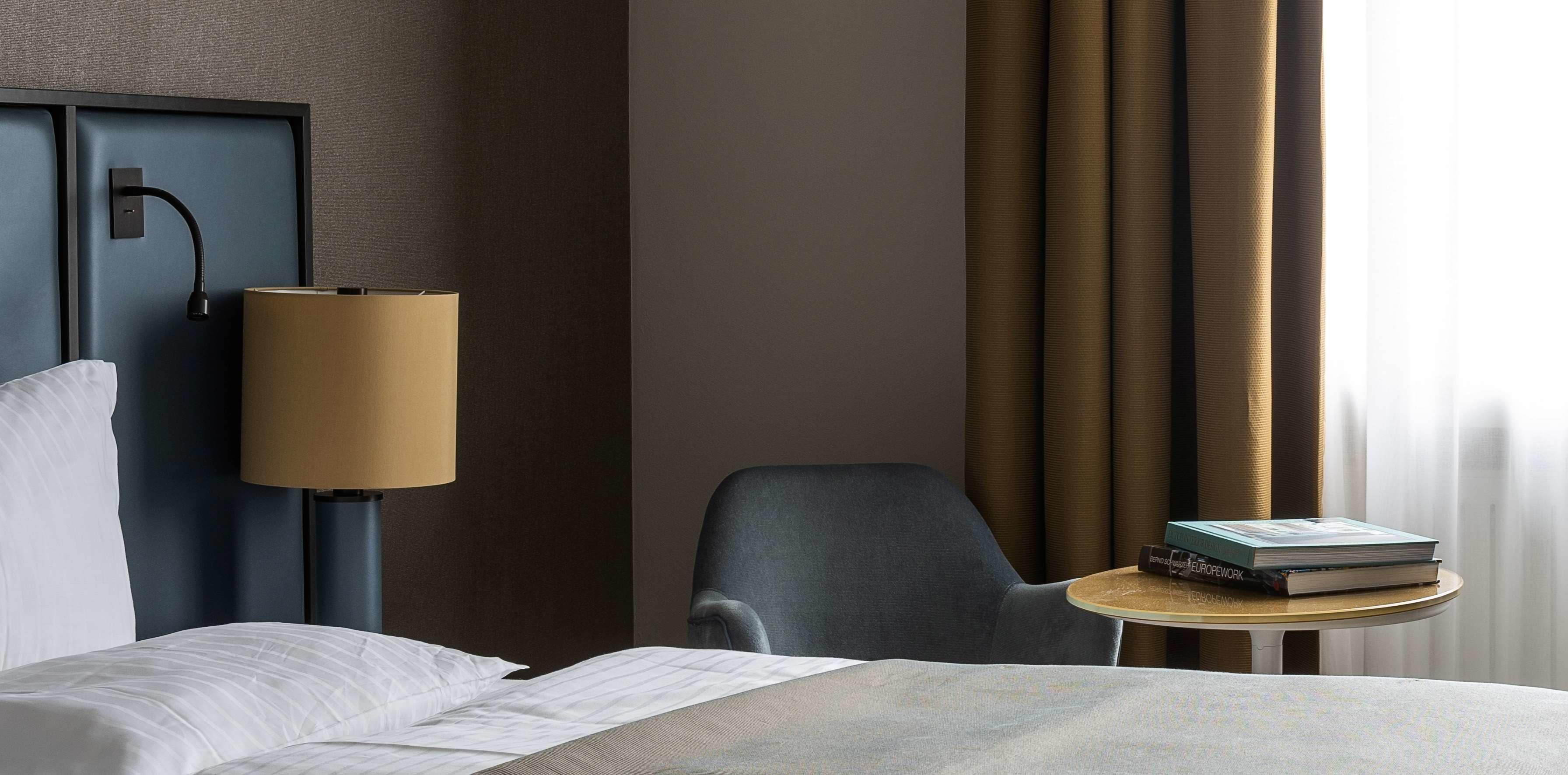 A hotel room with a bed, a desk with a lamp, a chair, and a window with curtains.
