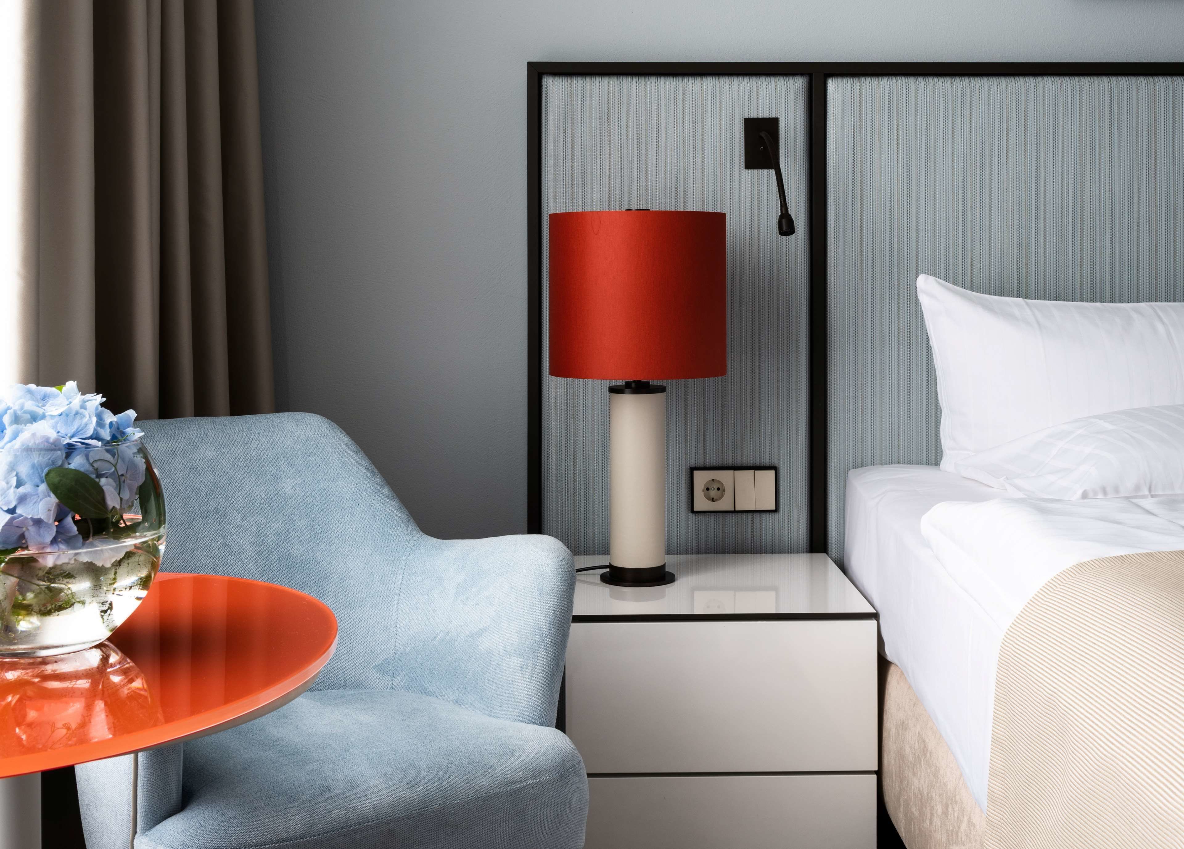 Een hotelkamer met een bed, nachtkastje, lamp, stoel en een tafel met een bloevas.