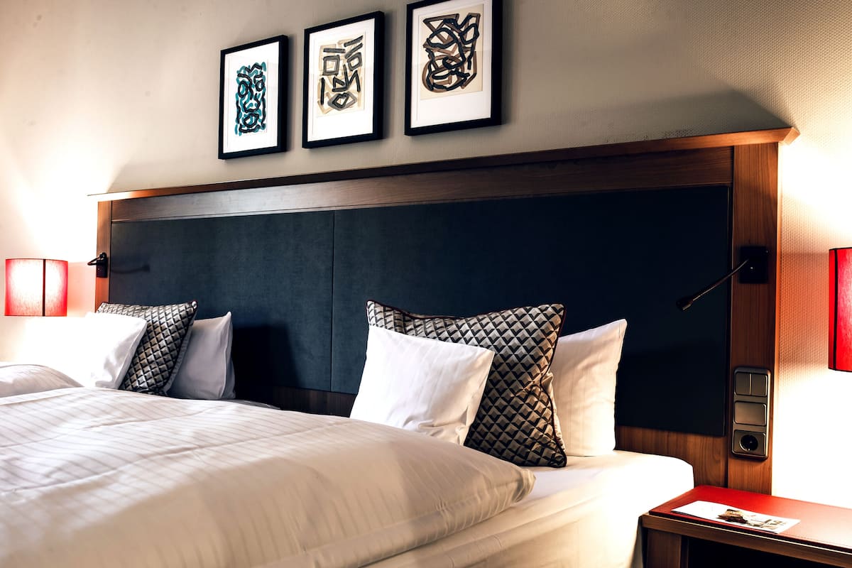 Un lit bien fait, avec du linge de lit blanc et des coussins à motifs, une tête de lit en bois et des œuvres d’art encadrées accrochées au mur.