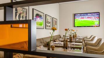 Un comedor con una televisión mostrando un partido de fútbol, un sofá y una mesa servida con vasos y flores.