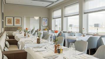 Un restaurante con largas mesas dispuestas para comer, obras de arte enmarcadas en las paredes y amplias ventanas con cortinas.