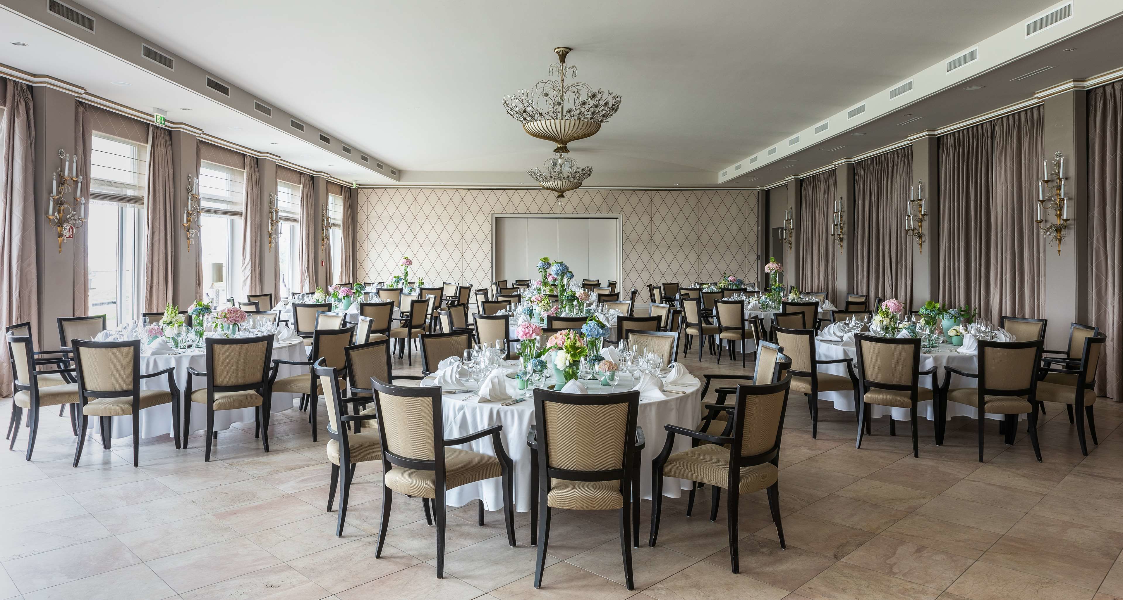A banquet hall with round tables set for dining, featuring chandeliers and wall-mounted sconces.