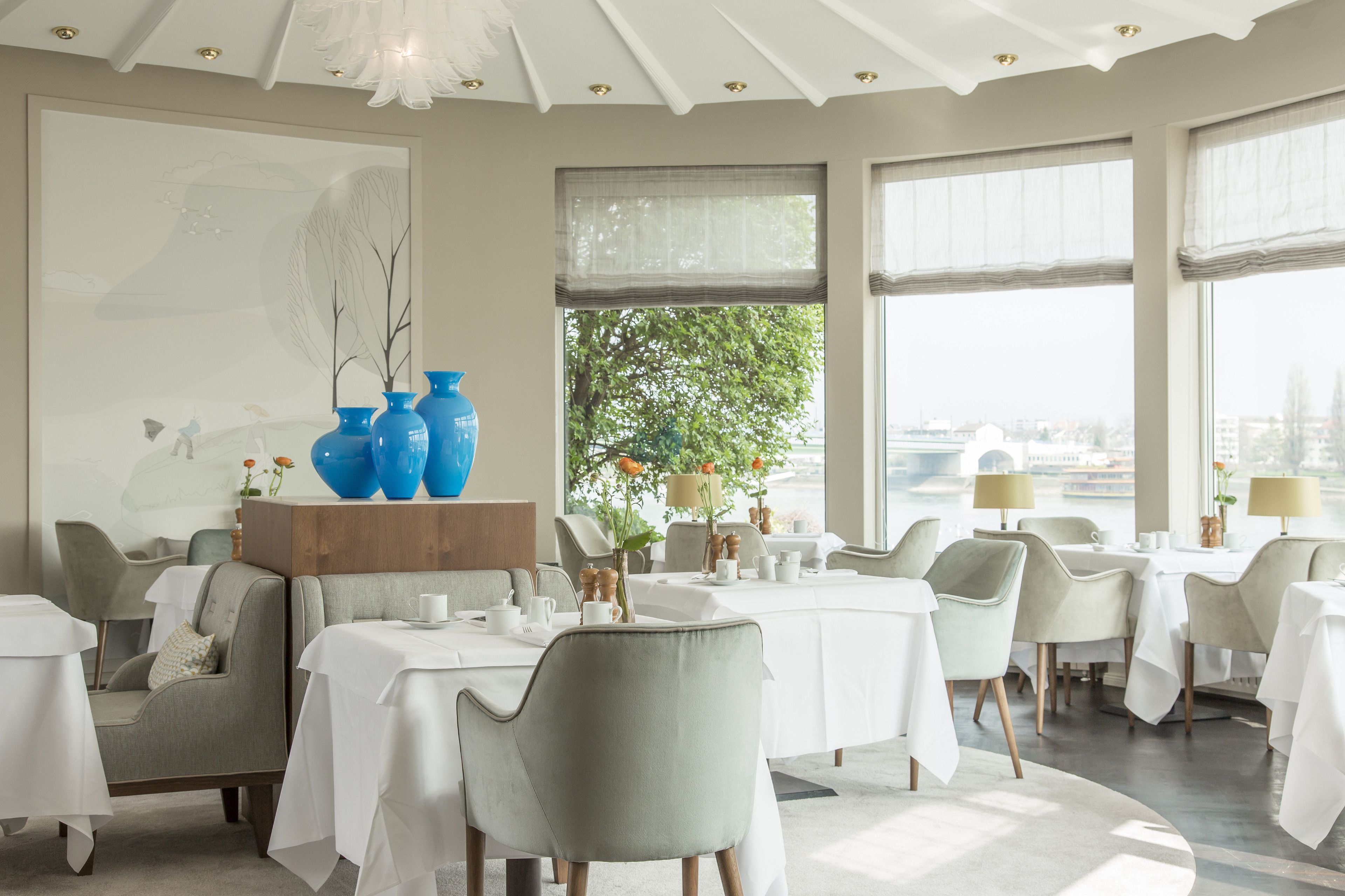 A dining area with round tables, grey upholstered chairs, and a large window with a view of trees and buildings.