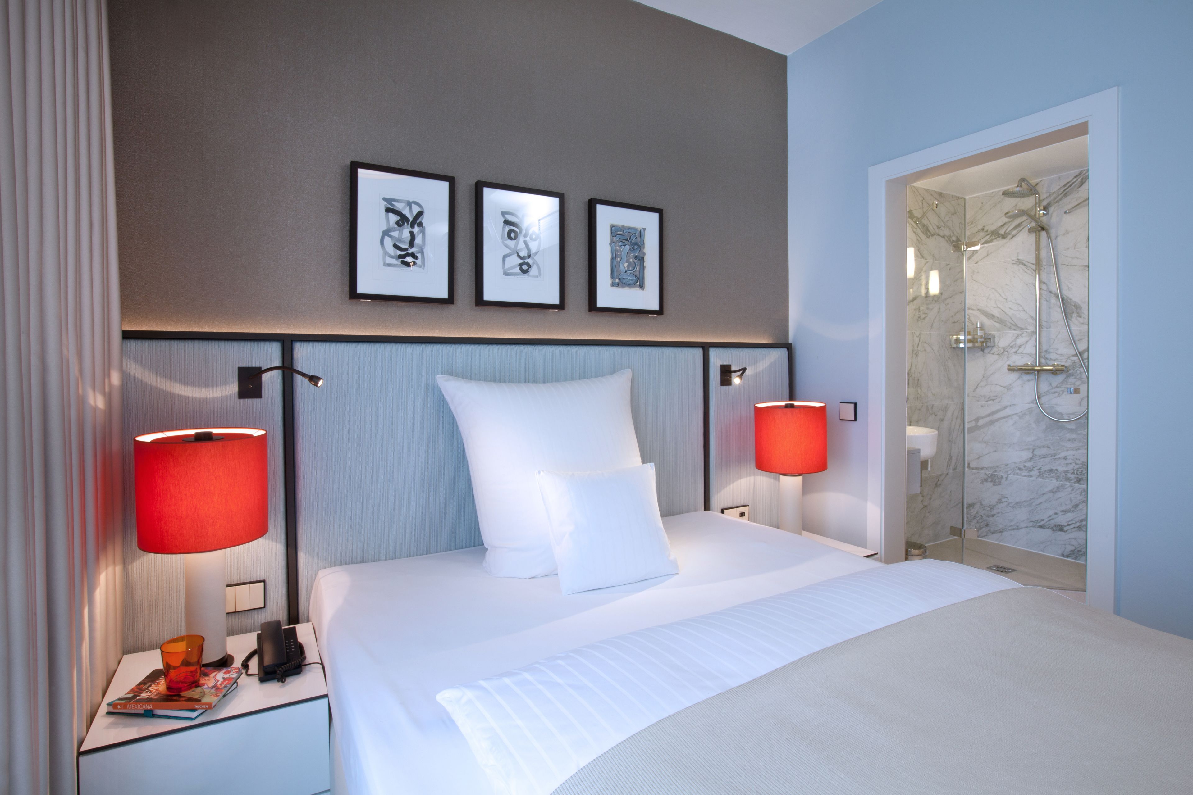 Une chambre d’hôtel dotée d’un grand lit, de deux lampes de chevet, d’une table de chevet avec un téléphone et un magazine, et d’un espace douche aux murs en marbre.